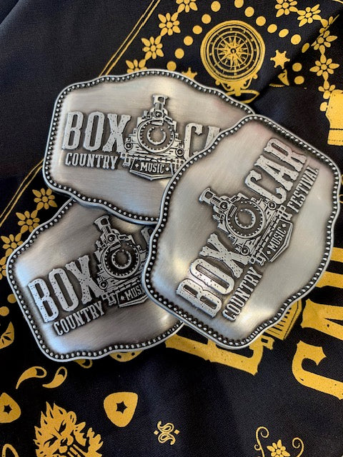 Three silver belt buckles with the text 'BOXCAR COUNTRY FESTIVAL' engraved on them, displayed against a backdrop with a yellow and black pattern.