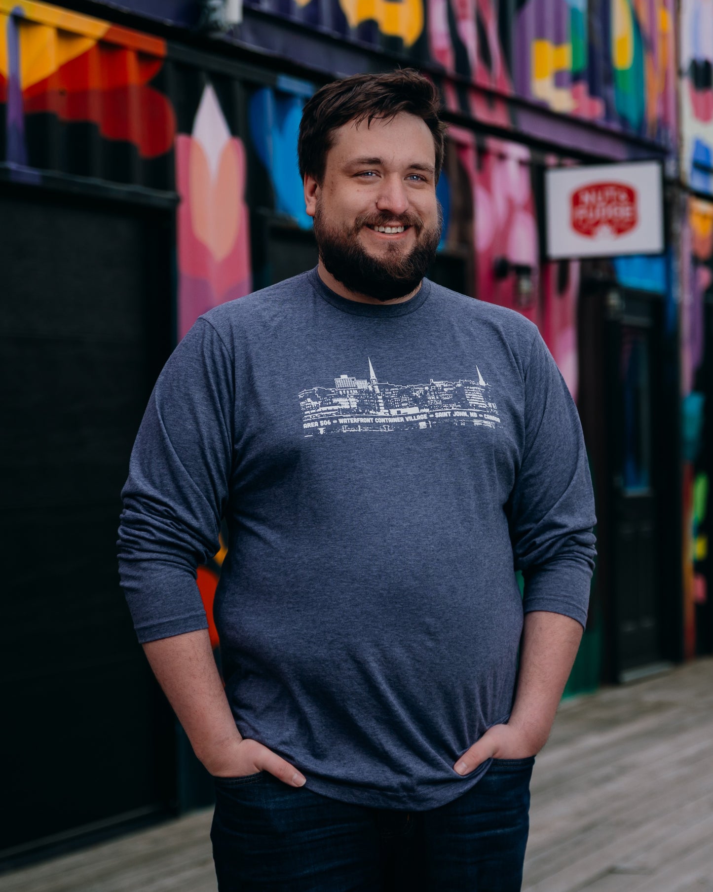Man wearing a blue shirt with a white line drawing, standing in front of a colourful mural.