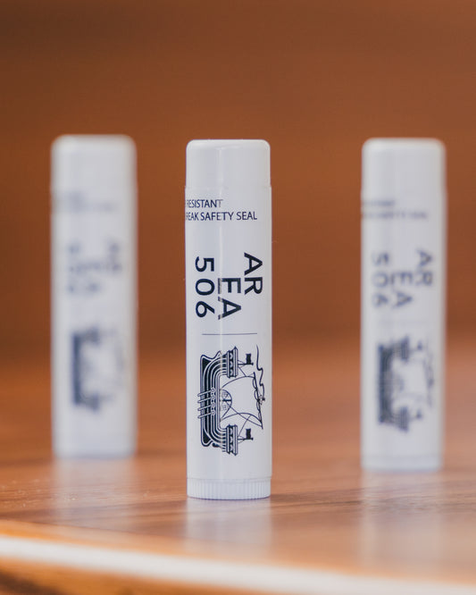 Three cylindrical AREA 506 lip balms on a wooden surface with a blurred background.