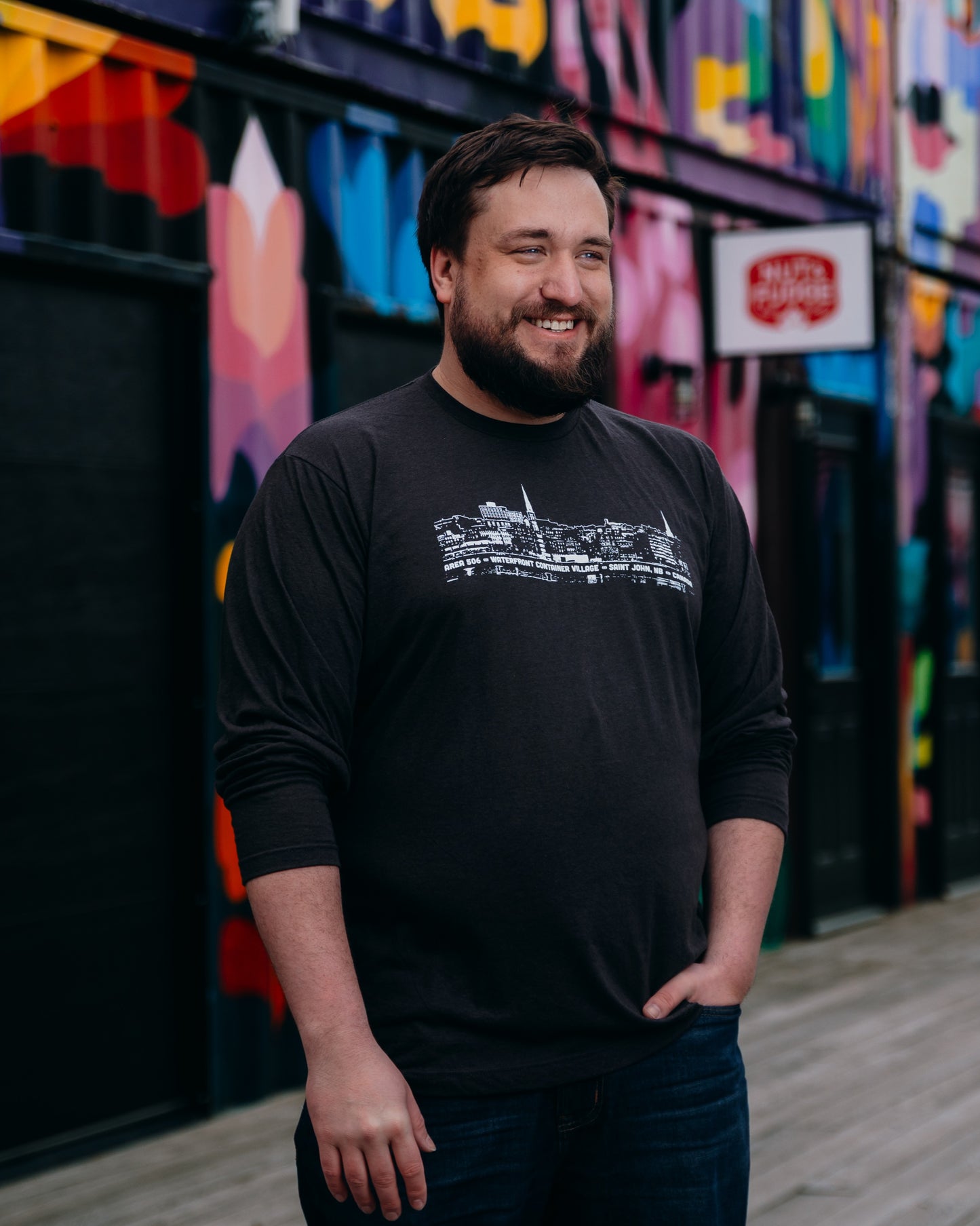Man wearing a black t-shirt with white line drawing, standing in front of a colourful mural.