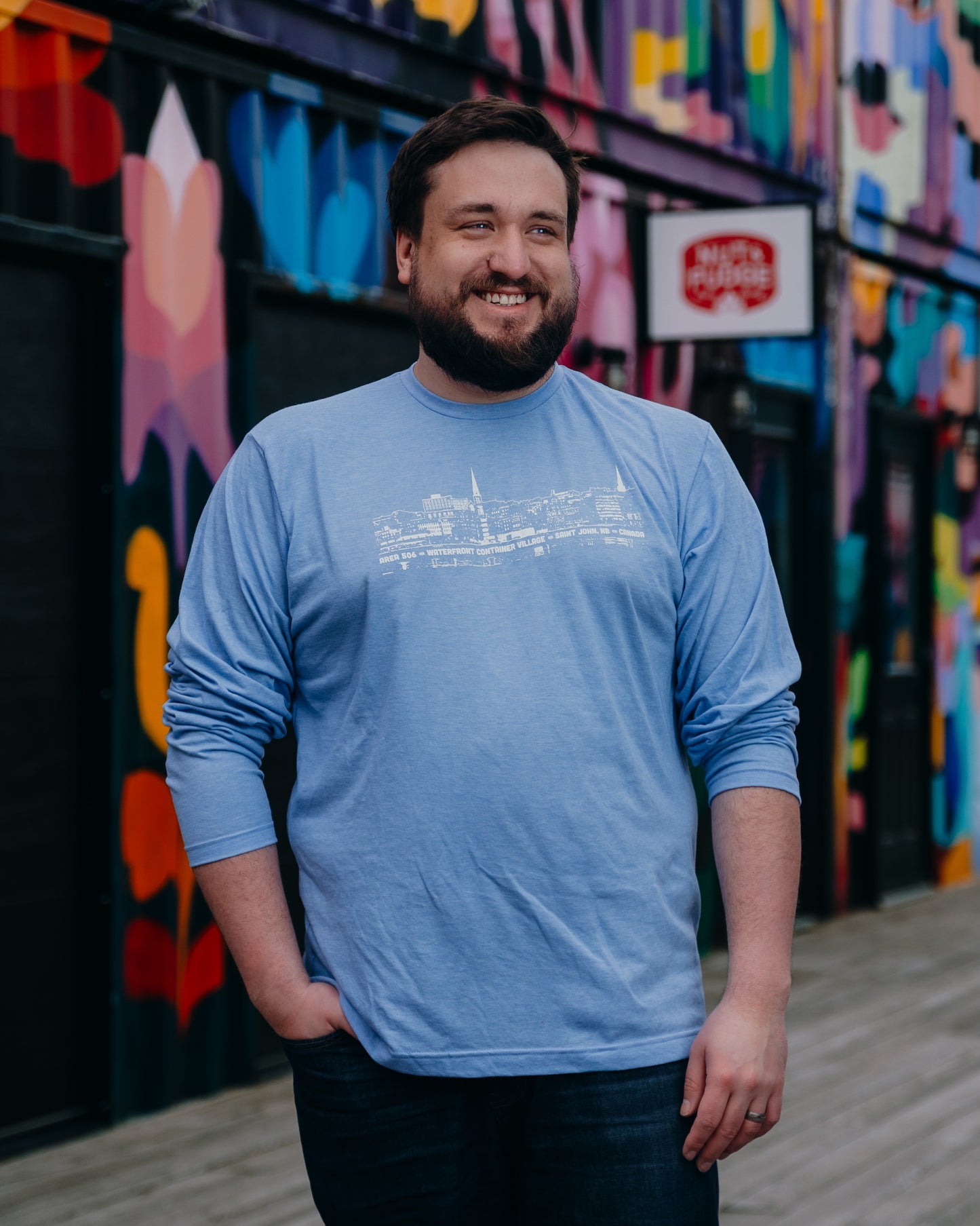 Man wearing a blue shirt with white line drawing, standing in front of a colourful mural.