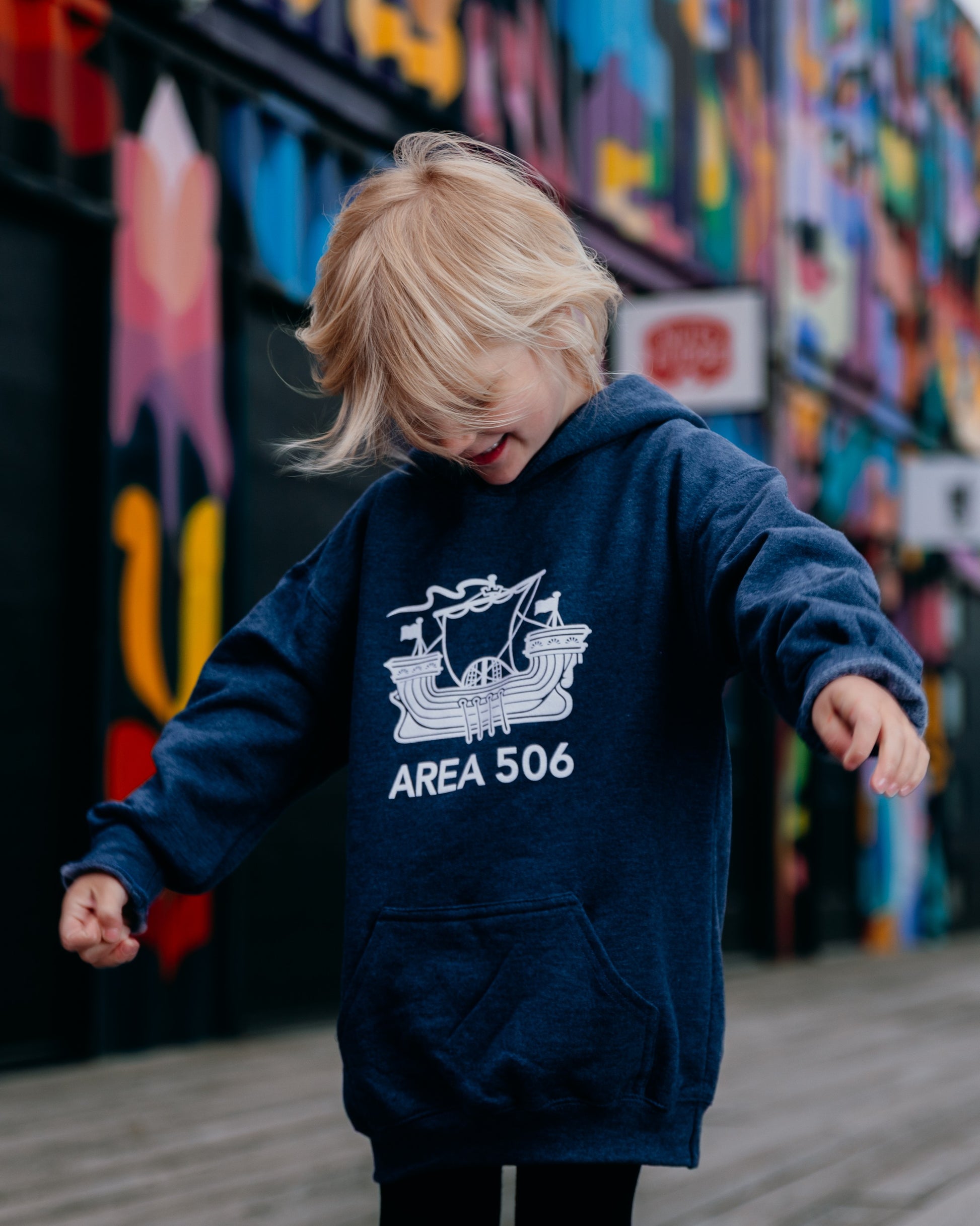 Child wearing a navy hoodie with 'AREA 506' design in front of a colourful mural.