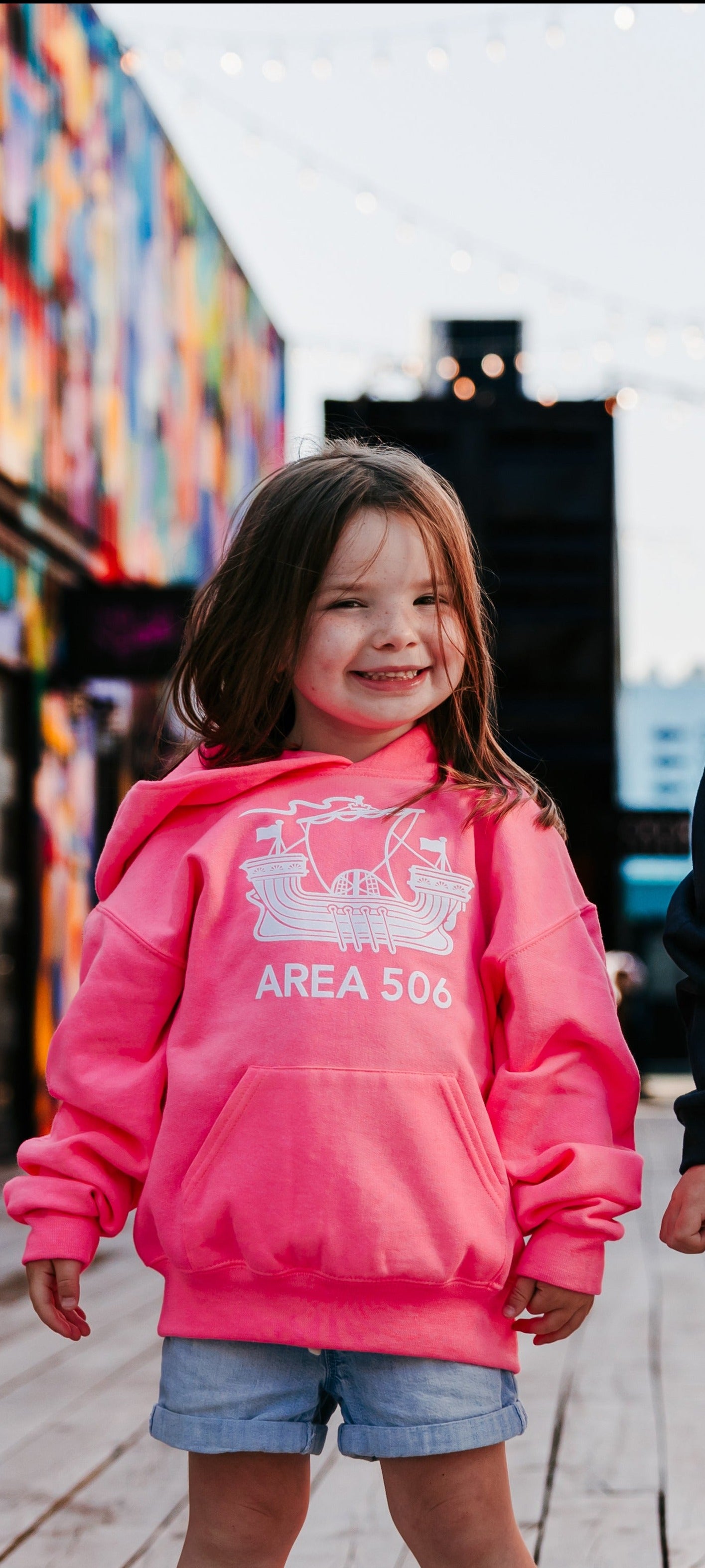 Child wearing a pink hoodie with 'AREA 506' design in front of a colourful mural.